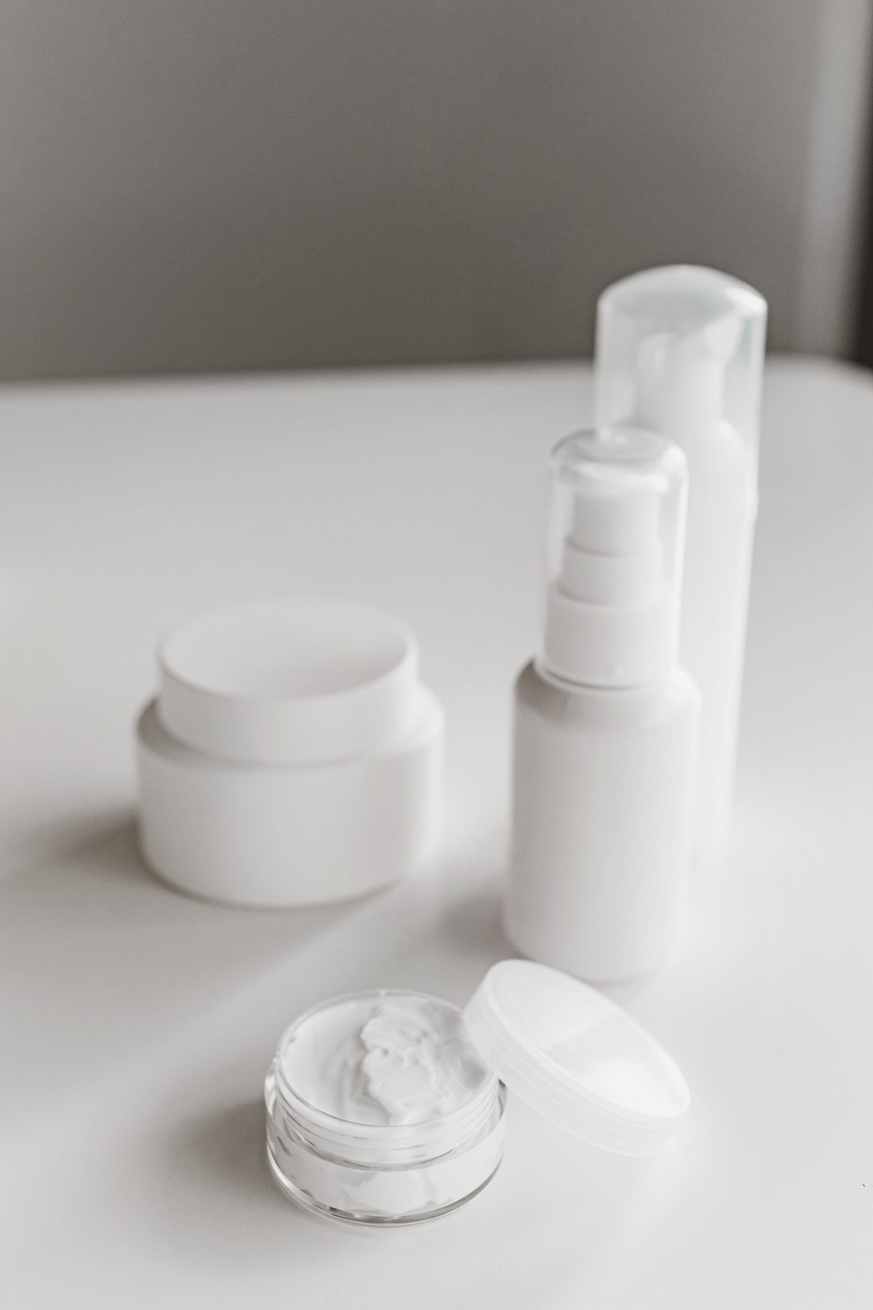White skincare jars and pump bottles arranged on a light surface, with an open jar of cream in the foreground.